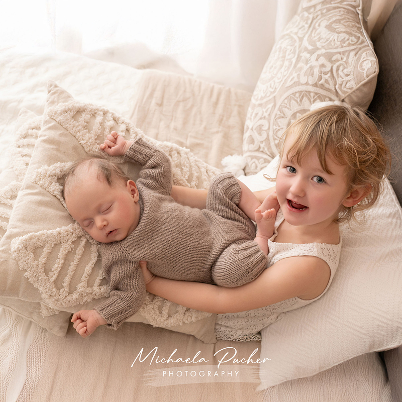 Ein kleines Mädchen liegt auf weichen, beigen Kissen und hält liebevoll ihr schlafendes Neugeborenes, das in einem gestrickten Outfit liegt.